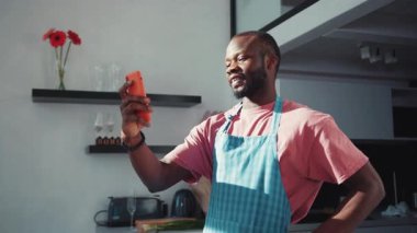 Çekici Afrikalı önlüklü adam akıllı telefon kullanarak mutfakta arkadaşıyla görüntülü konuşma yapıyor. Sosyal uzaklık. Çevrimiçi iletişim.