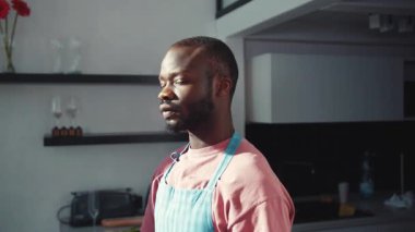 Modern mutfakta mavi yemek önlüğü giymiş, ciddi bir Afro-Amerikan erkeği duruyor. Yüz portresi. Yakışıklılar. Daire. Kapalı.