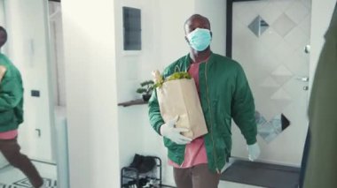 Yemek alışverişinden sonra eve dönen yakışıklı bir adam. Genç Afrikalı bir adam eve giriyor ve maskesini çıkarıyor. Karantina sırasında sağlık sigortası. Coronavirüs.