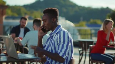Yakın plan yakışıklı Afro-Amerikalı genç adam dizüstü bilgisayarla masada oturmuş açık havada burger yerken teknoloji çok ırklı interneti yavaş çekimde kullanıyor.