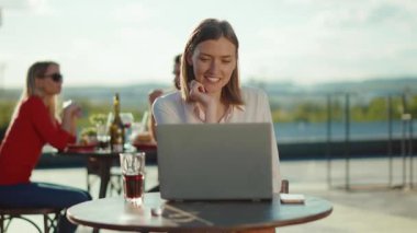 Gülücükler saçan genç bir kadın kafeteryada dizüstü bilgisayarla oturmuş internette internet haberleşme teknolojisi yazarak dışarıda ağır çekim iş yapıyor.