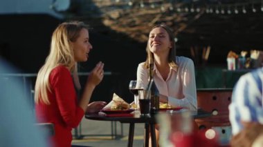 Dinlenen iki kadın telefonda konuşuyor. Kokteyl içiyorlar. Yazlık cafe caddesinde patates kızartması yiyorlar.