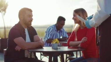 Gün ışığında mutlu genç bir çift parkın dışındaki masada oturmuş yemek siparişi veriyor. Yemek siparişi veriyor. Hafta sonları çalışan adam kafenin önünde perakende satış siparişi satıyor.