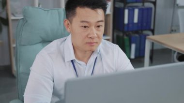 Ofisteki bilgisayar ekranına bakan ciddi bir iş adamının portresi. Asyalı erkek yönetici klavyeye basıyor, kameraya bakıyor ve gülümsüyor..