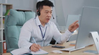 İşadamı kablosuz kulaklık takıp bilgisayarda konferans videosu çekiyor. Yetenekli, mutlu pazarlamacı müşterilere reklam yapıyor, notlar yazıyor..