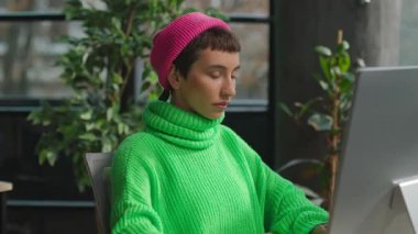 Açık yeşil kazaklı, ikili olmayan birinin iş yerinde kameraya baktığı bir fotoğraf.