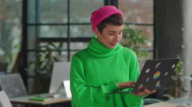 Elinde dizüstü bilgisayarla kameraya bakan açık yeşil kazaklı bir transseksüel.