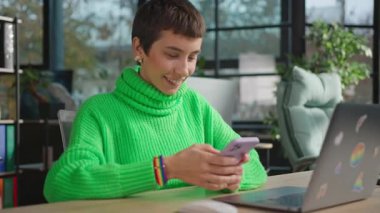 Gülümseyen genç eşcinsel kadın iş yerinde oturuyor ve akıllı telefondan içeride sohbet ediyor.