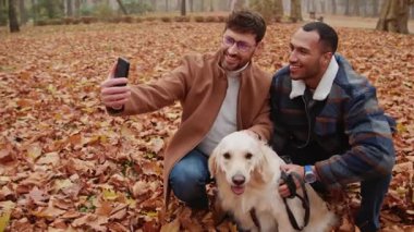 Açık havada köpekle selfie çeken gülümseyen gay erkeklerin portresi.