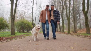 Gülümseyen, aynı cinsiyetten erkek, çok ırklı bir çift köpekle parkta yürüyor.