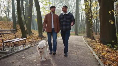 Romantik bir buluşma sırasında iki gülümseyen eşcinsel el ele tutuşup köpekle parkta yürür.