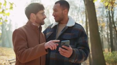 Gülümseyen, çoklu etnik kökenli erkekleri kucaklayan, akıllı telefon ekranına bakan,
