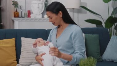 Yeni doğmuş bir bebekle oynayan ve modern bir dairede kanepede oturmuş gülümseyen mutlu bir annenin şirin portresi. Çocukluk. Annelik. Yavaş çekim