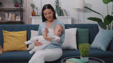 Evde mutlu genç bir anne kucağında yeni doğmuş bir bebekle oynuyor ve modern bir dairede koltukta oturmuş gülümsüyor. Çocukluk. Annelik. Yavaş çekim