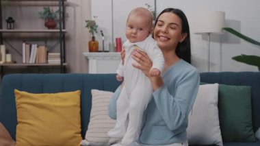 Yeni doğmuş bebekle oynayan ve modern bir dairede kanepede oturup gülümseyen sevimli, mutlu bir annenin portresi. Çocukluk. Annelik. Yavaş çekim