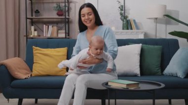 Yeni doğmuş bir bebekle oynayan ve modern bir dairede kanepede oturup gülümseyen mutlu bir annenin portresi. Çocukluk. Annelik. Yavaş çekim