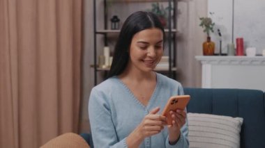 Mavi bir kanepede oturan ve akıllı telefon kullanarak modern bir odada gülümseyen kameraya bakan mutlu genç bir kadının portresi. Tıklıyor, kayıyor. Yavaş çekim