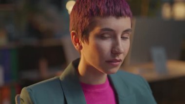 Pembe saçlı ve burnunda küpe olan genç bir kadın ofiste kameraya ciddi şekilde bakıyor.