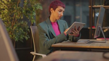 Gece ofiste pembe saçlı bir kadın oturuyor ve LGBT çıkartmalı tablet bilgisayar kullanıyor.