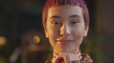 Pembe saçlı ve boğucu lgbt renklerinde şık bir kadın gece ofisindeki kameraya bakar.