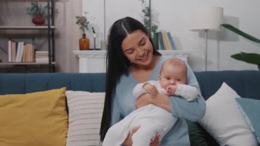 Gülümseyen genç bir anne yeni doğmuş bir bebeği kucağında tutuyor ve modern bir dairede bir kanepede oturuyor. Çocukluk. Annelik. Yavaş çekim