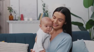 Mutlu genç anne portresi yeni doğmuş bebeğiyle oynuyor ve modern bir dairede bir kanepede oturuyor. Çocukluk. Annelik. Yavaş çekim