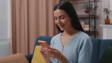 Mavi bir kanepede oturan çekici genç bir kadın ve modern bir odada etrafa bakıp gülümseyen bir akıllı telefon kullanıyor. Tıklıyor, kayıyor. Yavaş çekim