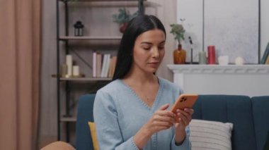 Güzel genç bir bayanı mavi bir kanepede oturtup, kafasını çevirerek modern bir odada kameraya bakan bir akıllı telefon kullanarak kapatın. Tıklıyor, kayıyor. Yavaş çekim