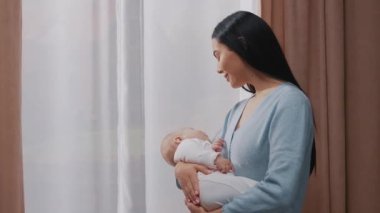 Mutlu anne yeni doğmuş bir bebeği kucağında tutuyor ve evdeki pencerelerin yanında duruyor. Gülümsüyor. Annelik. Çocukluk. Ebeveyn. Yavaş çekim