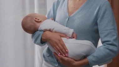 Gülümseyen bir anne yeni doğmuş bir bebeği kucağında tutuyor ve evdeki pencerelerin yanında duruyor. Gülümsüyor. Annelik. Çocukluk. Ebeveyn. Yavaş çekim