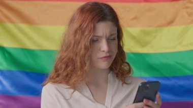 Üzgün genç bir kadın arka planda gökkuşağı bayrağı sallarken telefon gerginliği kullanır. LGBT
