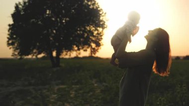 Annenin ve güzel bebeğin siluetleri. Anne kucağında bebekle tarlada öpüşüyor. Arka planda eski ağacın bulanık görüntüsü. Yaz zamanı, akşam vakti. İnanılmaz bir gün batımı. Açık hava.