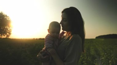Genç, güzel anne sükunetini sağlayan çocuğun görüntüsü. Bebeği elinde tutan muhteşem bir kadının fotoğrafı. Şefkat, sevgi, ilgi. Açık havada, şehir dışında. Gün batımı. Alanın bulanık arkaplanı. Kırsal alan