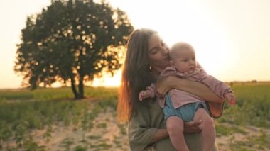 Yeşil, zarif elbisesi olan, bebeği elinde tutan, güzel çocuğu öpen beyaz bir kadın. Çekici anne kameraya bakıyor. Akşam vakti. Arka planda güzel bir doğa. Yaz zamanı, açık hava