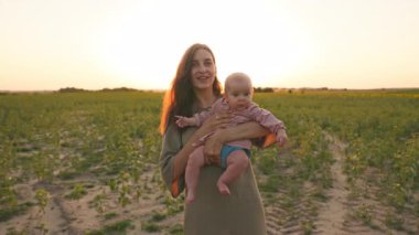 Genç, gülümseyen beyaz bir kadının portresi. Küçük, güzel bir kızı elinde tutuyor. Kameraya bakıyor ve gün batımında el sallıyor. Mutlu annelikler. Kırsal alan. Açık havada. Yaz. Aşk