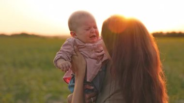 Genç anne ve ağlayan kızın tarladaki portresi. Beyaz bir kadının kucağında sevimli bir bebekle çocuğu öptüğü görüntüler. Dışarıda, şehir dışında. Gün batımında akşam. Şefkat
