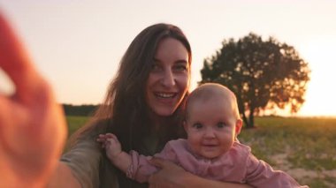 Güzel görünümlü beyaz bir kadın güzel bir bebeği elinde tutuyor, akıllı telefonda özçekim yapıyor, doğadaki kameraya bakıyor. Kız ve bebek gülümsüyor. Anne ve çocuk kavramı. Açık havada. Saha. Gün batımı