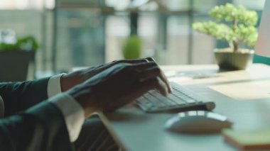 İş yerinde oturan ve klavyeye yazı yazan, tanınmayan Afrikalı orta yaşlı bir ofis çalışanının erkek ellerine yakın çekim. Bonsai 'nin bulanık görüntüsü ve bilgisayar monitörü. Kapalı