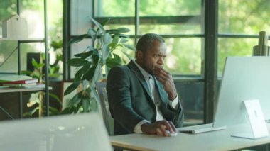 İş yerinde oturan ciddi bir Afrikalı adamın vurulması, proje hakkında önemli bilgilerin olduğu e-postaları okumak. Bilgisayar kullanırken heyecanla gülümseyen iş adamının portresi. Başarı, keyif konsepti. Kapalı