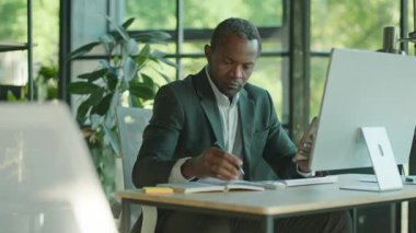 Bilgisayarla masaüstünde oturan, elinde akıllı telefon tutan, defterine yazı yazan ciddi odaklı Afro-Amerikan bir adamın portresi. Gri takım elbiseli yakışıklı iş adamının fotoğrafı. İçeride, ofiste, aletlerde.