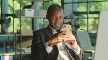 Orta yaşlı Afrikalı bir adamın iş yerinde oturup akıllı telefondaki bir kadınla sohbet ettiği görüntüler. Gülümseyen zeki, güzel sakallı iş adamının portresi. İletişim, internet. Modern rahat ofisin yeşil bulanık arka planı