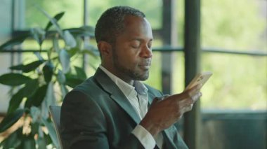 Bilgisayarın başında oturan ve iş ortağına sesli mesaj kaydeden yakışıklı, orta yaşlı, kendine güvenen Afrikalı bir adamın yatay çekimi. Ofis çalışanı elinde akıllı telefon, el kol hareketi. İçeride. Gündüz, teknoloji.