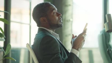 Gri takım elbiseli, akıllı telefon kullanarak iş yerinde oturan düşünceli, orta yaşlı Afrikalı bir adamın portresi. İş ortaklarına sesli mesaj kaydeden kendine güvenen iş adamlarının görüntüleri. İçeride. Açık alan, ofis
