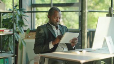 Resmi giysiler içinde bilgisayarda oturan ve tablet üzerinde gezinen Afrikalı bir adamın görüntüsü. Gülümseyen başarılı iş adamının portresi. İçeride. Ofis