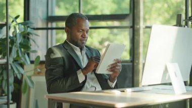 Gri takım elbiseli, güzel sakallı, gülümseyen Afrikalı bir adamın bilgisayarda oturup tableti elinde tutuşu. Alet kullanan bir iş adamı. Cihaz, elektronik, modern teknoloji. İçeride, gündüz vakti, rahat bir ofiste.