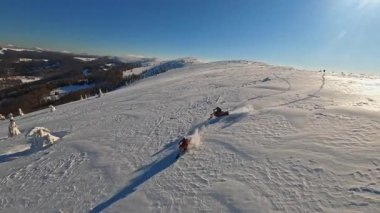 Kar motosikletli iki adam kışın güneş ışığında atlayıp gezerler. Dağlarda gökyüzü görünümü