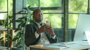 Yakışıklı Afrikalı Amerikalı adam internet üzerinden iletişim kurmak için akıllı telefon mesajları kullanıyor. Rahatına bak. İş yerindeki modern güneş ışığı ofisinde oturmuş gülümsüyorum. Modern aygıtlar, işyeri