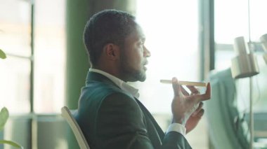 Takım elbiseli yakışıklı Afro-Amerikan iş adamı iş görüşmeleri yaparken ya da ofiste ses kaydederken mutlu ve gülümserken. Güneş ışığı. Sohbet. Yavaş çekim