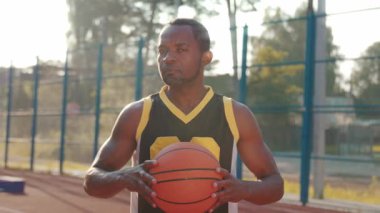 Çekici Afro-Amerikan basketbolcunun basketbol sahasında topuyla dikilirken çekilmiş yatay görüntüleri. Kameraya bakan sağlıklı bir adamın portresi. Spor konsepti, aktiviteler, yaz zamanı. Bulanık arkaplan