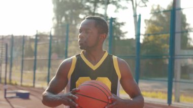 Basketbol sahasında kameraya gülümseyen yakışıklı Afrikalı Amerikalı basketbol oyuncusunun portresi. Güneşli bir günde arka planı bulanık duran mutlu adam. Aktif yaşam tarzı, açık hava.